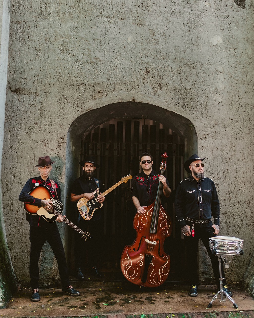 Os quatro integrantes do Farol Negro em frente ao arco do forte, com contrabaixo acústico e caixa de bateria